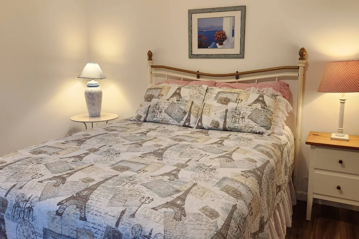 A serene bedroom with an Eiffel Tower patterned bedspread, soft table lamps on either side, and framed artwork adorning the walls. Natural morning light fills the room, enhancing its comfortable and restful atmosphere.