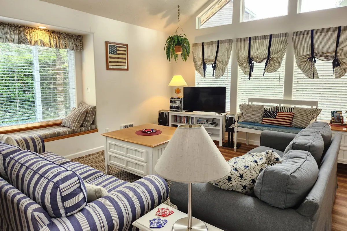 The hero image shows a bright, inviting living room featuring a striped sofa, a cozy window seat perfect for reading or gazing outside, a central coffee table ideal for games or snacks, and a TV for shared movie nights. Tasteful patriotic accents add a cheerful, Americana vibe to the space, making it feel warm and welcoming for families or groups of friends.