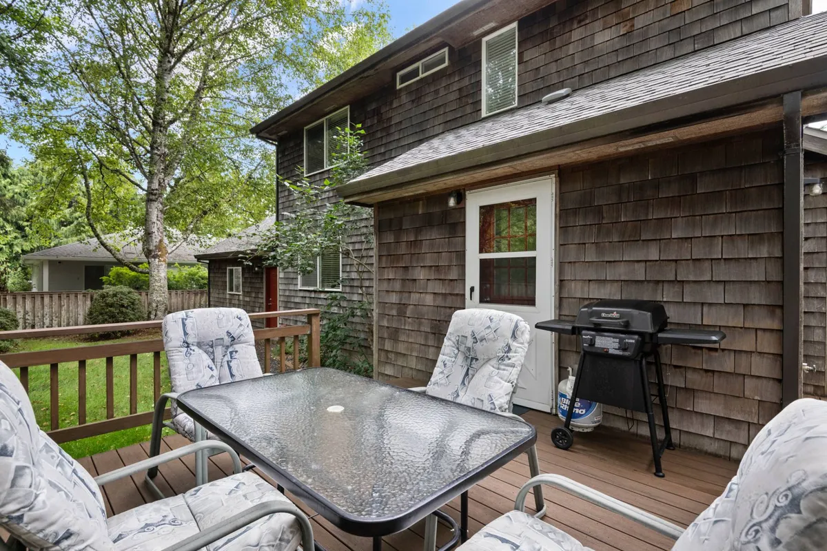 An outdoor deck scene featuring a glass table with comfortable chairs and a barbecue grill, surrounded by lush greenery. The mood is casual and relaxed, perfect for outdoor dining and social gatherings during warm, sunny days.