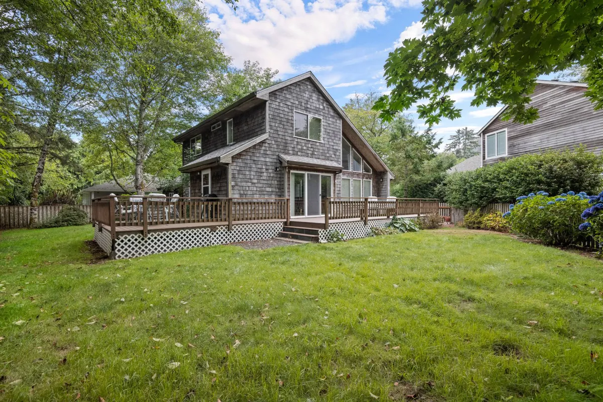 This image showcases a welcoming exterior with a wooden deck overlooking a lush green lawn, detailed wooden shingles, and mature trees surrounding the yard. The open, sunny backyard is fully fenced, offering a safe play area for children and pets, while the deck provides space for outdoor dining and relaxation.