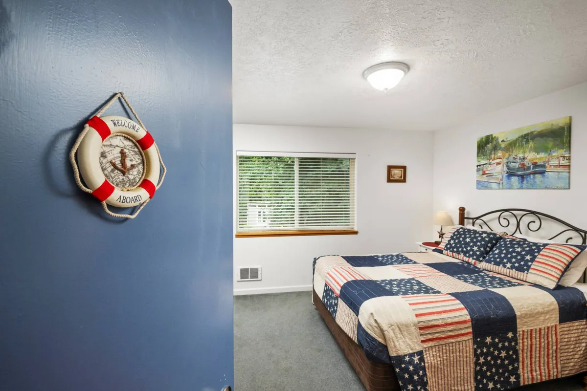 A cozy bedroom adorned with a nautical-themed quilt, a 'welcome aboard' sign on the door, and a landscape painting. Soft lighting and an inviting bed create a warm ambiance, ideal for unwinding after a day at the beach.
