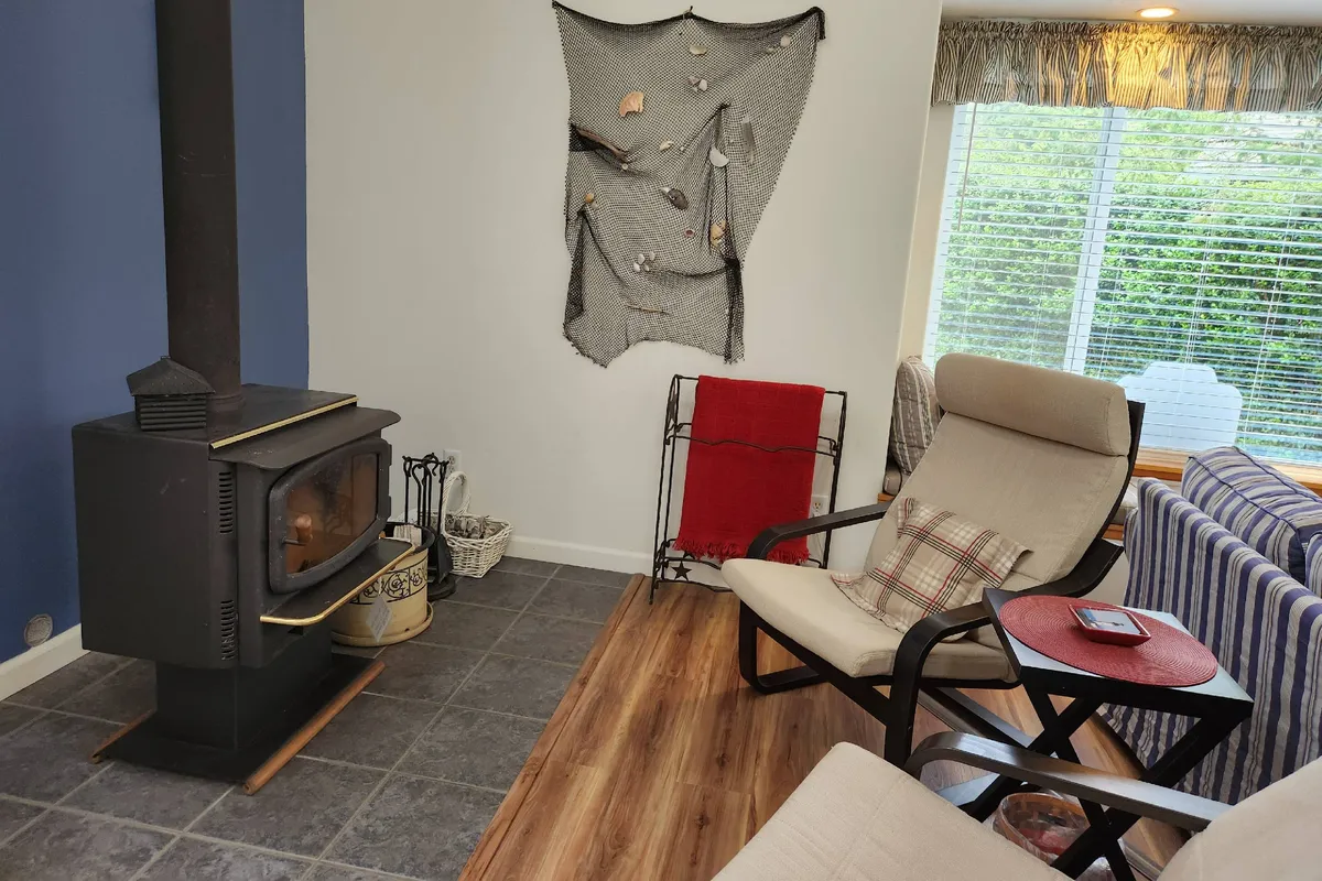 An inviting corner of the living room showcasing a wood stove, comfortable armchairs, a textile wall hanging, and a window letting in soft, natural light. The ambiance is perfect for relaxing by the fire or enjoying intimate conversations on rainy coastal days.