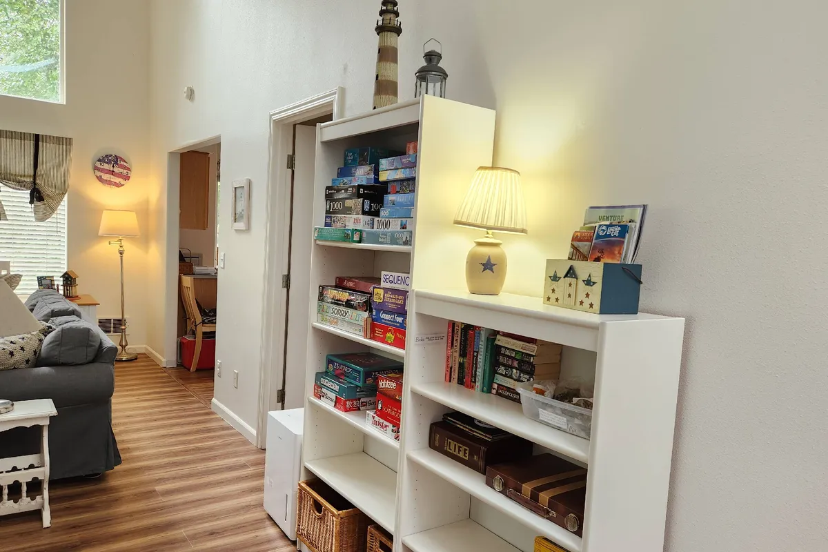 A warmly lit living space featuring bookshelves filled with games and books, a cozy lamp, and a nautical lighthouse decor piece. The scene evokes a sense of comfort, perfect for family game nights or unwinding with a book after a day of coastal adventures.