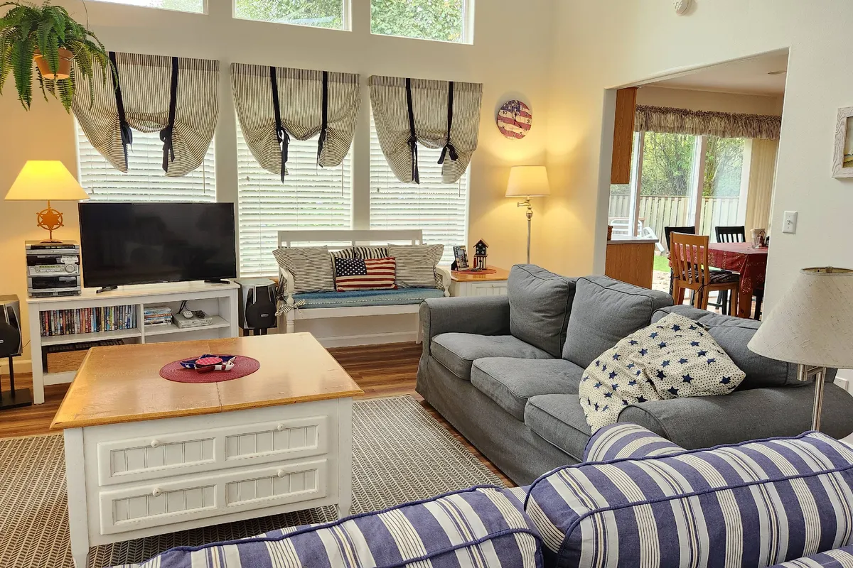 A sunlit living room with striped couches, a wooden coffee table, a TV unit with media, and decorative cushions. The space is arranged for socializing, watching movies, or enjoying quiet moments, all within a bright and welcoming setting.
