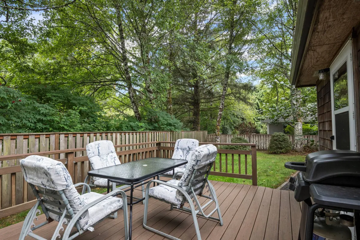 An exterior image of a backyard with a dining table, chairs, grill, abundant surrounding greenery, and a visible fence. The setup invites outdoor dining, barbecues, and relaxed gatherings, clearly showing the safety and versatility of the yard for families and pets.