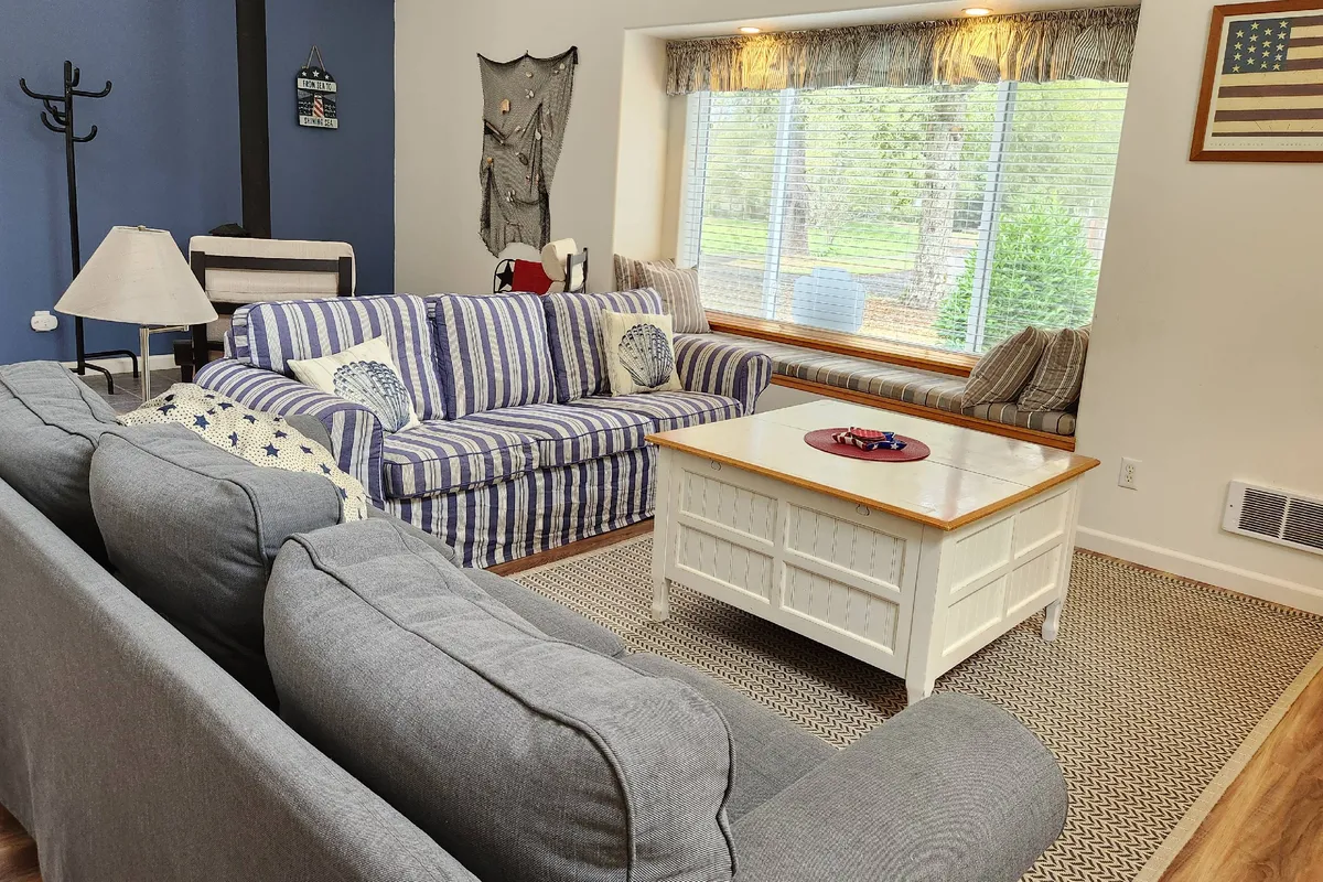 This image features a striped sofa and a white coffee table positioned near a large window, through which ample natural light pours in. The window creates a visual connection to the outdoors, suggesting the presence of beautiful surroundings just outside. The relaxed setup invites guests to unwind and reflects a seamless indoor-outdoor experience.